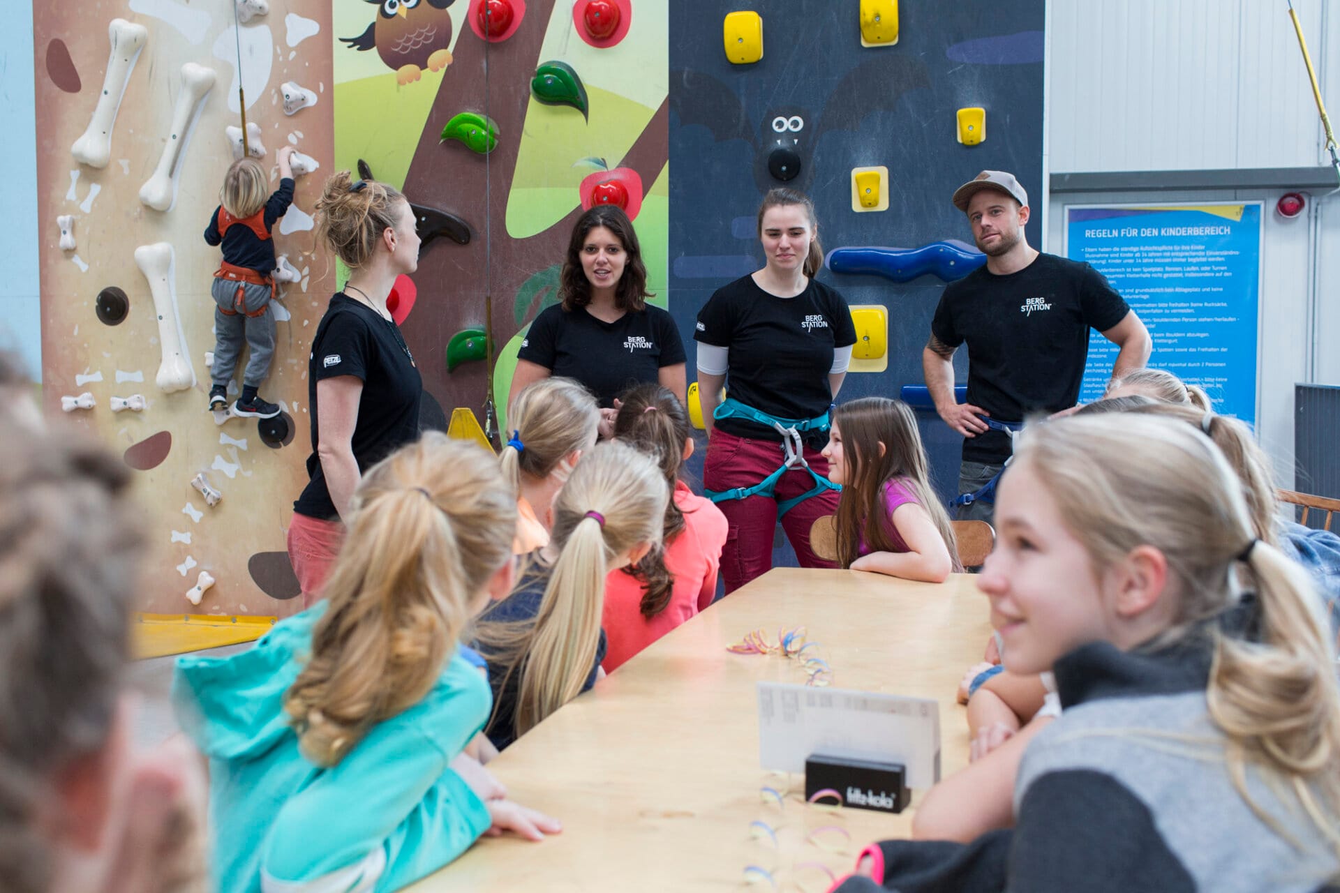 Trainer*innen im Hintergrund und Kindern im Vordergrund vor einer Kletterwand